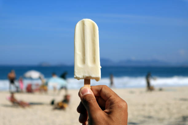 Man's hand holding a coconut ice cream bar against blurry seaside beach