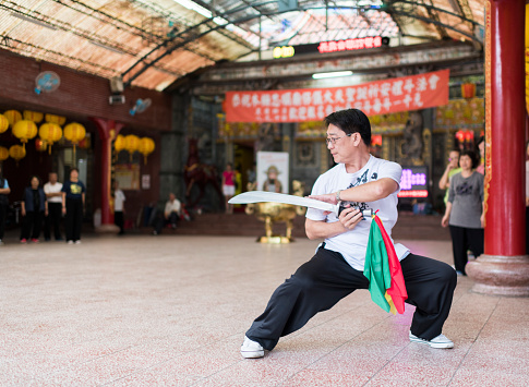 Wide portrait of a man practicing Wushu in a temple courtyard with a traditional sword.
