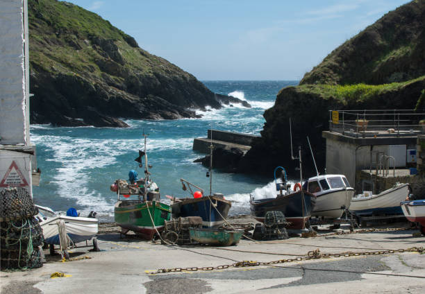 Row of fishing boats on slip way in Portloe harbour, Cornwall with harbour wall and crashing waves in the background.