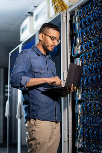 Concentrated network engineer examining database server Concentrated handsome young Arabian bearded network engineer in glasses standing by network server cabinet and examining errors on laptop database photos stock pictures, royalty-free photos & images