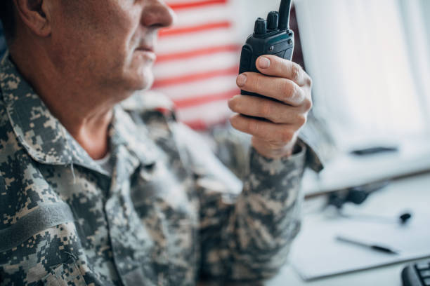 One senior US soldier in uniform sitting at the office and using walkie- talkie with American flag on the wall.