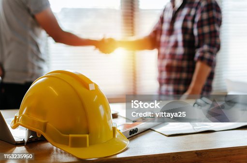 istock yellow safety helmet on workplace desk with construction worker team hands shaking greeting start up plan new project contract in office center at construction site, partnership and contractor concept 1153235950