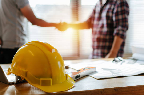 yellow safety helmet on workplace desk with construction worker team hands shaking greeting start up plan new project contract in office center at construction site, partnership and contractor concept - engineer architect building contractor team imagens e fotografias de stock