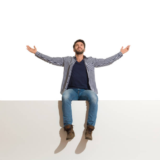 Young man in boots, jeans and unbuttoned lumberjack shirt is sitting on a top with arms outstretched and looking up. Full length studio shot isolated on white.