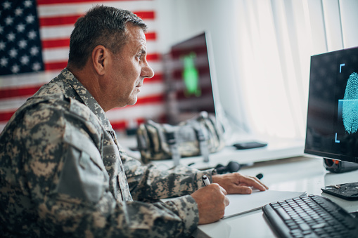 One senior soldier in uniform investigating fingerprints on computer and doing paperwork at the office with American flag on the wall.