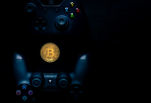 SHEFFIELD, UK - JUNE 2ND 2019: Overhead shot of a Microsoft black Xbox one controller and A Sony black Playstation 4 controller with a physical gold bitcoin in the middle of them both against a black background