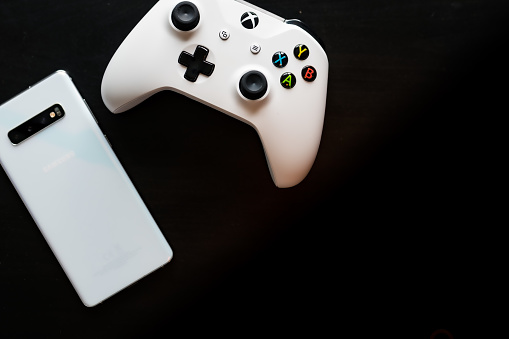 Shot taken from above against a dark wooden background showing a White Samsung Galaxy S10+ and a Xbox One White Controller next to each other with room for text on the right hand side
