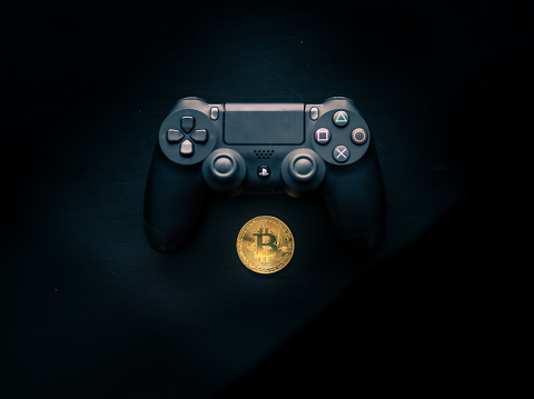 SHEFFIELD, UK - JUNE 2ND 2019: Shot taken from above of a Black Sony Playstation 4 controller sat above a Physical gold bitcoin on a dark black background