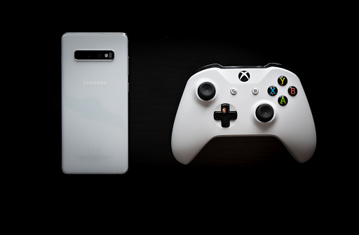 Shot taken from above against a dark wooden background showing a White Samsung Galaxy S10+ and a Xbox One White Controller next to each other in the middle of the screen to represent multi platform gaming