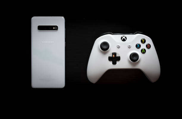 A phone and controller sit side by side as gaming becomes multi-platform Shot taken from above against a dark wooden background showing a White Samsung Galaxy S10+ and a Xbox One White Controller next to each other in the middle of the screen to represent multi platform gaming brand name games console stock pictures, royalty-free photos & images