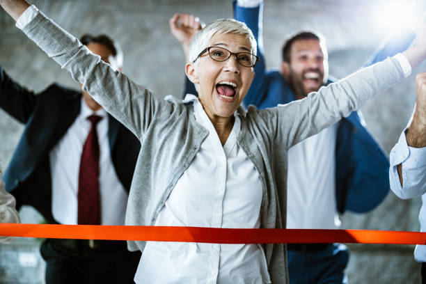 Cheerful senior businesswoman celebrating her success while crossing the finish line in the office. Happy mature businesswoman crossing finish line during sports race in the office. race office stock pictures, royalty-free photos & images