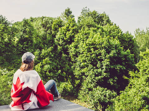 Cute woman and Canadian Flag on a background of trees and blue sky. View from the back, close-up. National holiday concept