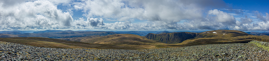An panorama view of a Scottish mountain summit plateau with heather and cliff under a majestic blue sky and huge white clouds