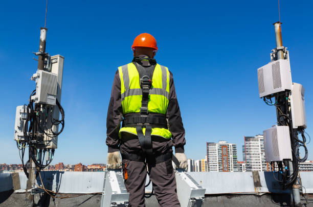 istock Telecommunication engineer  in helmet and uniform holds telecomunication equipment in his hand and  antennas of GSM  DCS UMTS LTE bands, outdoor radio units, optic fibers, power cables are installed on the roof. Working process of upgrading telecommunicat 1153266570