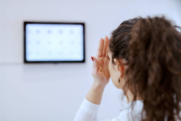 Brunette with curly hair and ponytail looking at chart test vision. Optician shop interior.