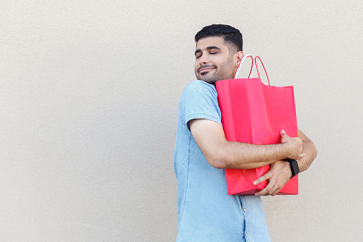 The best gift ever. Portrait of handsome young bearded man in blue shirt standing, hugging his lovely red shopping bag or gift and enjoying. indoor studio shot isolated on light beige wall background.