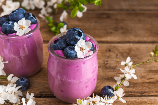 Two glasses of blueberry yogurt with fresh blueberries and spring flowers on rustic wooden background. close up