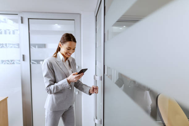 belle femme d’affaires caucasienne dans l’usure formelle et avec les longs cheveux bruns utilisant le téléphone intelligent et entrant au bureau. - women computer home interior brown hair photos et images de collection