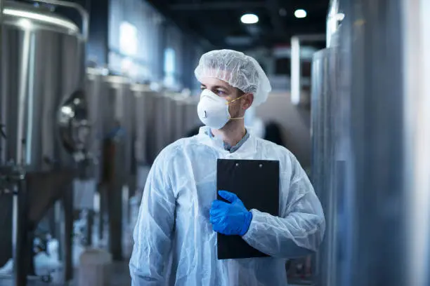 Photo of Technologist in protective white suit with hairnet and mask standing in food factory.
