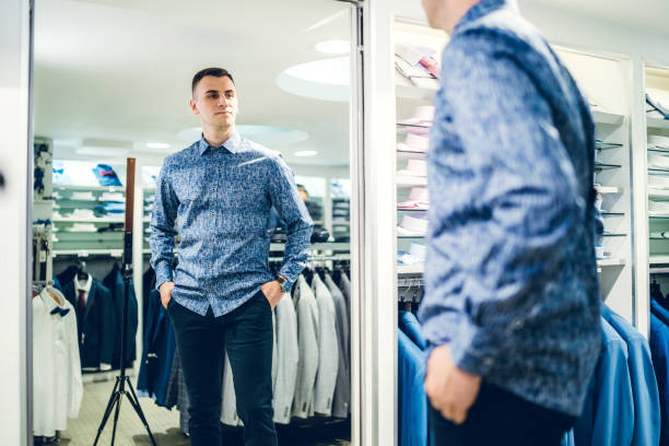 Man trying shirt in a store Young handsome caucasian man trying shirt in a store and looking himself in a mirror. fitting room stock pictures, royalty-free photos & images