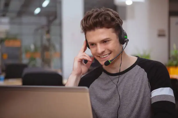 Young handsome man working at call center, wearing headset with microphone, copy space. Friendly male customer support operator answering callers, typing on laptop