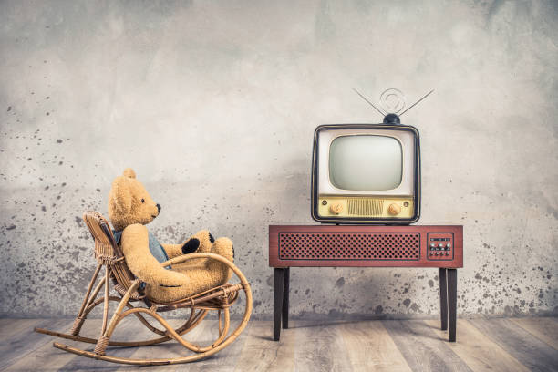 Retro Teddy Bear toy on rocking chair watching old outdated analog television receiver from 50s on wooden TV stand front aged loft textured concrete wall background. Vintage style filtered photo