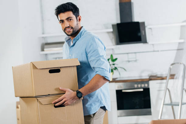 cheerful latin man holding boxes while moving in new home stock photo