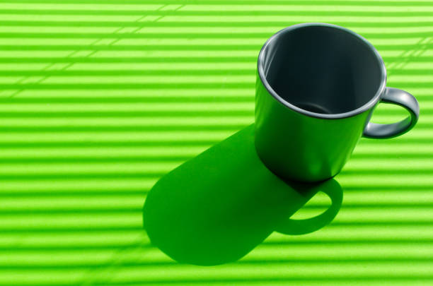 Grey mug on a green background with hard shadows from the rays of the bright sun making its way through the blinds.