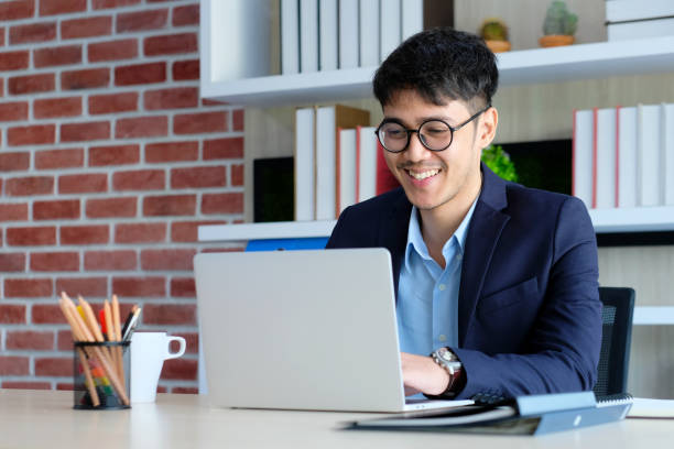 istock Young asian businessman smiling while working with laptop computer at office, business office lifestyle concept 1153312979