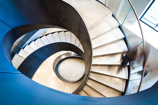 Wide angle color image depicting an abstract high angle view of a spiral staircase. We can see the blurred motion of a businessman walking down the spiral staircase, giving the impression that he is moving fast.  Room for copy space. ***IMAGE WAS SHOT IN CITY HALL, LONDON, UK, A PUBLICLY OWNED BUILDING FREELY ACCESSIBLE TO THE PUBLIC. THERE ARE NO FEES OR TICKET PURCHASES NECESSARY TO ENTER OR PHOTOGRAPH THIS BUILDING, NOR ARE THERE ANY PHOTOGRAPHIC RESTRICTIONS***