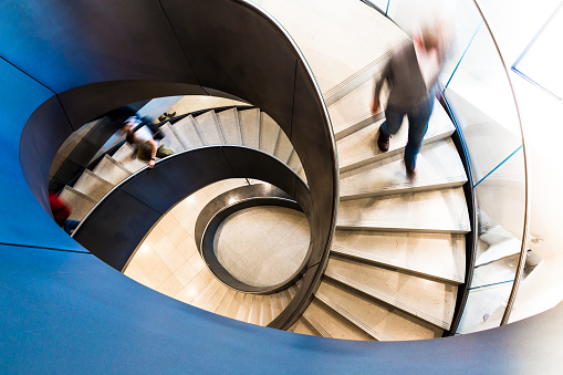 Overhead view depicting blurred motion of people walking up and down a spiral staircase. The people are completely blurred, and the slow shutter speed makes it appear as though the people are moving fast. Image taken at City Hall in London, which is a publicly owned building that is accessible to the general public without any photographic restrictions. Room for copy space.