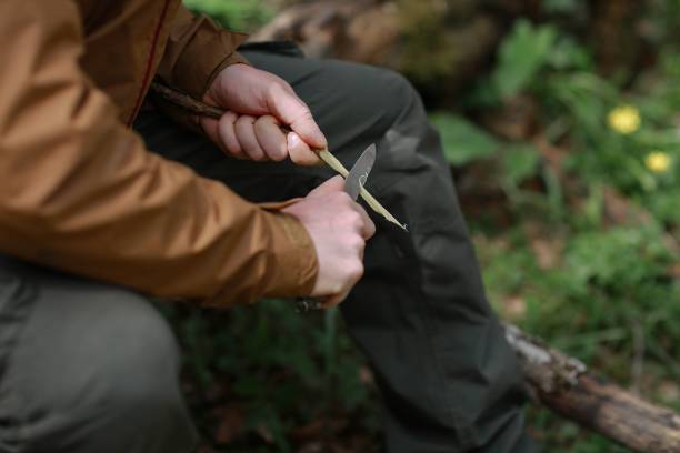 Man sharpens a branch with a knife Person in a brown windbreaker sharpens a branch with a knife in the Crimean forest. Travel, adventure, hiking and motivation concept. craft knife stock pictures, royalty-free photos & images