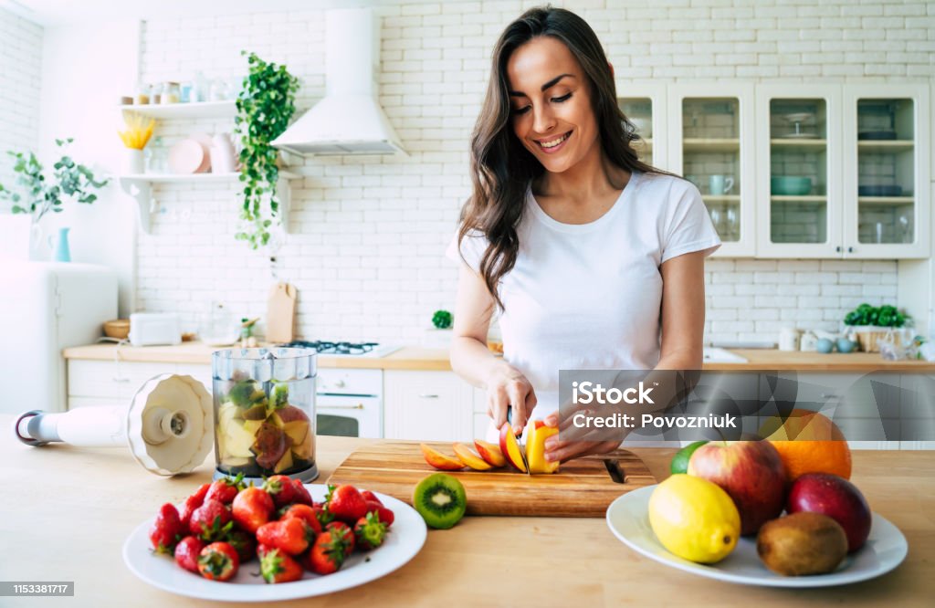 Beautiful woman making fruits smoothies with blender. Healthy eating lifestyle concept portrait of beautiful young woman preparing drink with bananas, strawberry and kiwi at home in kitchen. Healthy Eating Stock Photo