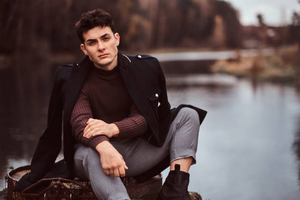 istock A handsome man with a coat covering his shoulders looking at camera while sitting near a lake in the autumn forest. 1153382005
