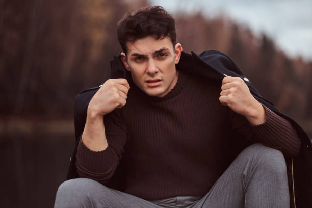 istock A handsome man with a coat covering his shoulders looking at camera while sitting near a lake in the autumn forest. 1153382051