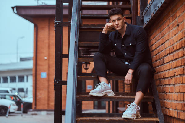 A young pensive guy sitting on stairs outside near a building with the industrial exterior. A young pensive guy wearing a shirt sitting on stairs outside near a building with the industrial exterior. urbane stock pictures, royalty-free photos & images