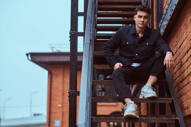 A fashionable guy sitting on the stairs outside the building with the industrial exterior. stock photo