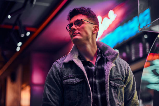 a young man fashionably dressed standing in the street at night. illuminated signboards, neon, lights. - neon light fotos imagens e fotografias de stock