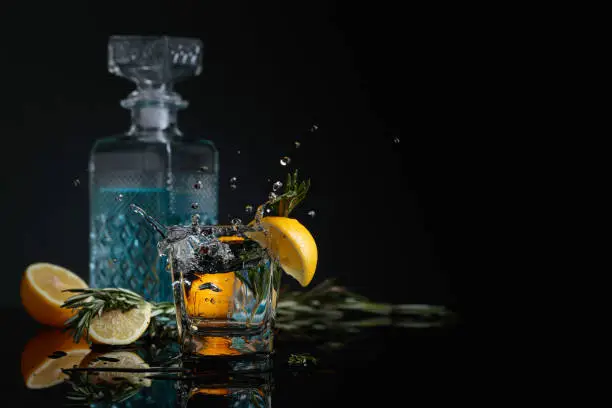 Gin-tonic with lemon slices and twigs of rosemary. Piece of ice falls into the glass. Copy space on a black background.
