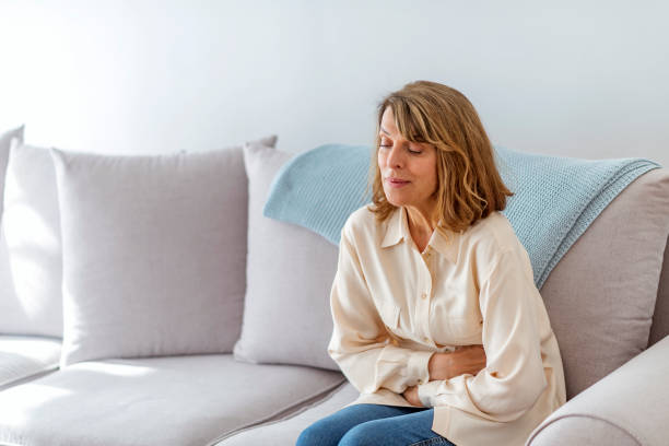 I wonder if the doctor does house calls Senior woman suffering from stomach ache sitting on a bed in the bedroom. Photo of Senior woman sitting on her bed has a stomach ache in the bedroom. Senior woman suffering from stomach pain while sitting on a gray sofa during the day. stomach cancer stock pictures, royalty-free photos & images