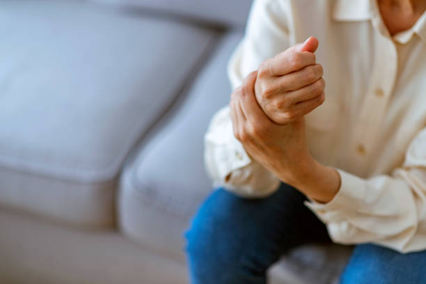 Woman holding her wrist symptomatic Photo of Painful Wrist In An Elderly Person. Senior woman suffering from pain in wrist at home while sitting on a gray sofa. Mature woman feeling wrist pain, injury problem, healthcare concept, sprain rheumatoid arthritis stock pictures, royalty-free photos & images