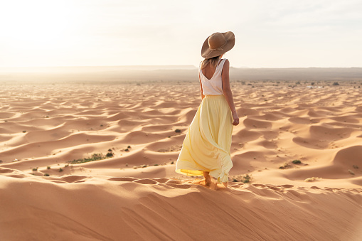 A young woman in a long yellow skirt, white shirt and straw hat is watching the dawn from the top of the sand dune of the Merzouga desert. Dawn in the Sahara desert.