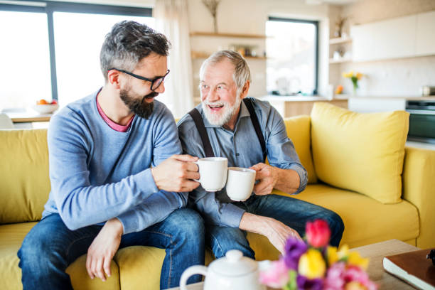 An adult son and senior father sitting on sofa indoors at home, drinking tea. An adult hipster son and senior father sitting on sofa indoors at home, drinking tea. elderly parents stock pictures, royalty-free photos & images