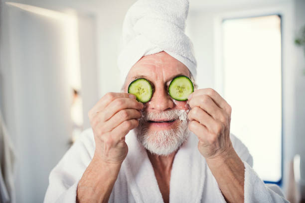 senior man with cucumber on front of his eyes in bathroom indoors at home. - facial mask spa treatment cucumber human face imagens e fotografias de stock