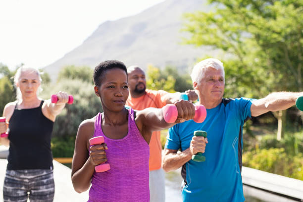 Mature fitness people exercising with dumbbells Happy senior and mature couples exercising with dumbbells. Healthy multiethnic people exercising using dumbbells outdoor. African couple and senior friends in sportswear stretching arms holding colorful dumbbells at park. black man white woman couple stock pictures, royalty-free photos & images