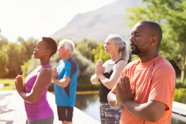 Mature people meditating together at park Four senior people meditating with joined hands and closed eyes breath deeply. Multiethnic group doing breathing exercise during yoga session outdoor. Class of mature people doing meditation with joined hands and relaxing together at park. meditation training stock pictures, royalty-free photos & images