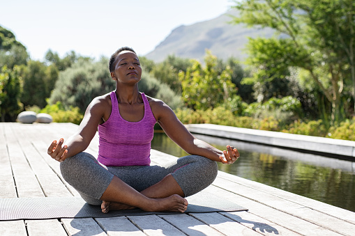 Mature african woman practicing yoga and meditates in lotus position near swimming pool outdoor. Fitness black lady sitting in lotus pose with closed eyes. African american woman meditating at poolside with copy space.