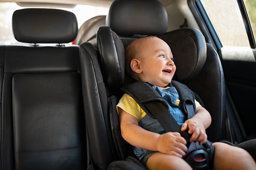 Portrait of cute smiling toddler sitting in car seat and looking outside the window. Little boy sitting in safety chair and laughing alone with copy spce. Happy child having fun in safety car seat while enjoy the travel.