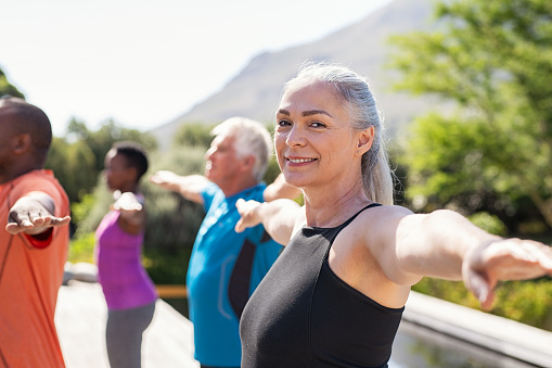 Portrait of happy senior woman practicing yoga outdoor with fitness class. Beautiful mature woman stretching her arms and looking at camera outdoor. Portrait of smiling serene lady with outstretched arms at park.