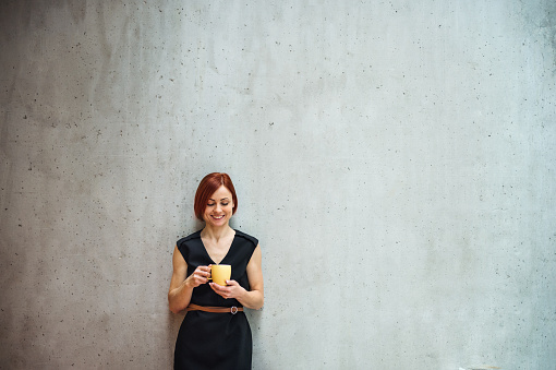 A portrait of young cheerful businesswoman standing in office, holding a cup of coffee.